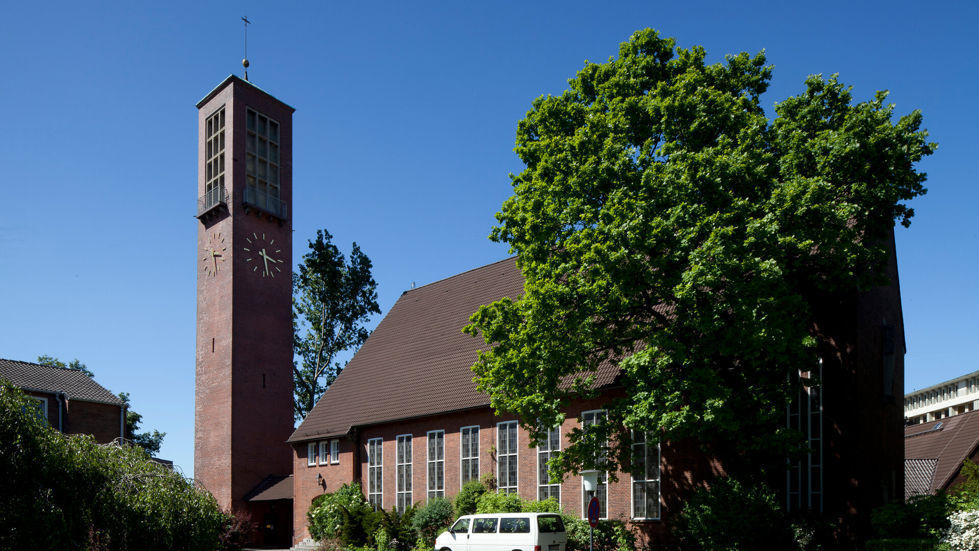 "Evangelischer Gottesdienst- Hier bin ich zuhause - Du auch": Die Erlöserkirche in Hamburg ist aus dunklem Backstein gebaut mit einem schlichten, eckigen Kirchturm. Sie ist von einigen Laubbäumen und Büschen umgeben, dahinter ist der Himmel blau.
