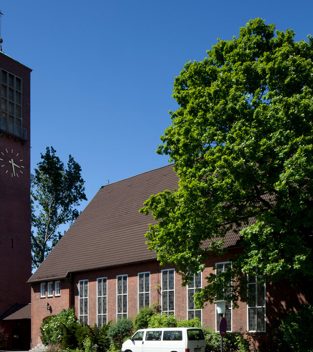 "Evangelischer Gottesdienst- Hier bin ich zuhause - Du auch": Die Erlöserkirche in Hamburg ist aus dunklem Backstein gebaut mit einem schlichten, eckigen Kirchturm. Sie ist von einigen Laubbäumen und Büschen umgeben, dahinter ist der Himmel blau.
