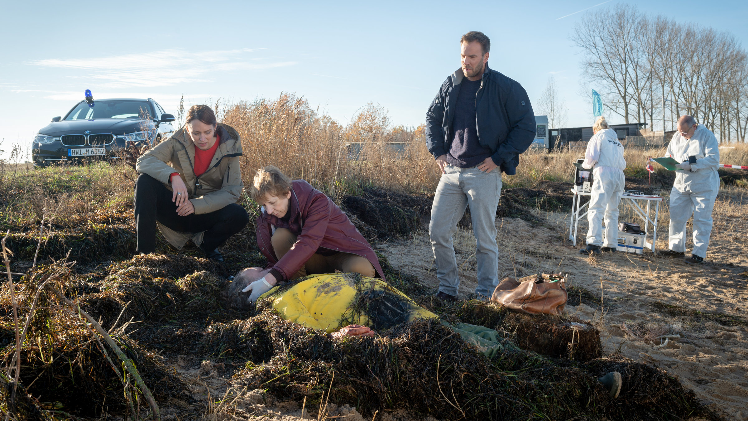 "SOKO Wismar - Strandpirat": Reinhard Koller (Michael Krämer) liegt leblos am Strand inmitten von angeschwemmten Algen. Helene (Katharina Blaschke) kniet neben ihm und tastet sein Genick ab. Neben ihr hockt Karoline Joost (Nike Fuhrmann). Pöhlmann (Dominic Boeer) steht neben der Leiche, die Hände in die Hüften gestemmt. Im Hintergrund sind zwei Kriminaltechniker. Karolines Wagen parkt am Straßenrand. Eine Baumreihe säumt die Straße.