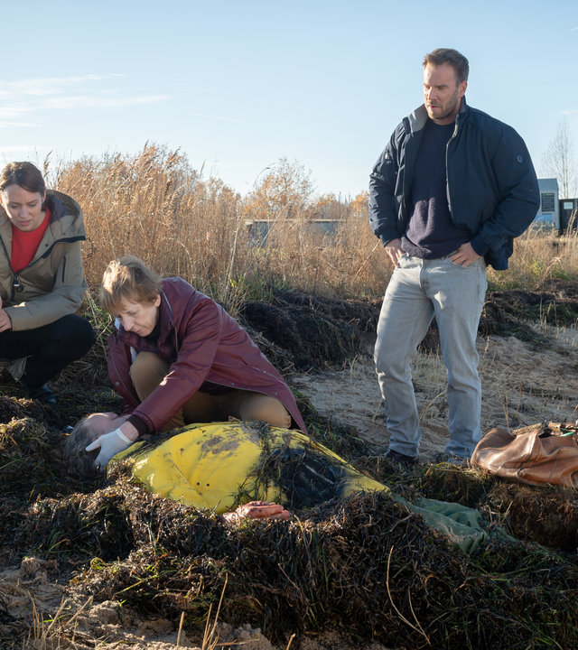 "SOKO Wismar - Strandpirat": Reinhard Koller (Michael Krämer) liegt leblos am Strand inmitten von angeschwemmten Algen. Helene (Katharina Blaschke) kniet neben ihm und tastet sein Genick ab. Neben ihr hockt Karoline Joost (Nike Fuhrmann). Pöhlmann (Dominic Boeer) steht neben der Leiche, die Hände in die Hüften gestemmt. Im Hintergrund sind zwei Kriminaltechniker. Karolines Wagen parkt am Straßenrand. Eine Baumreihe säumt die Straße.