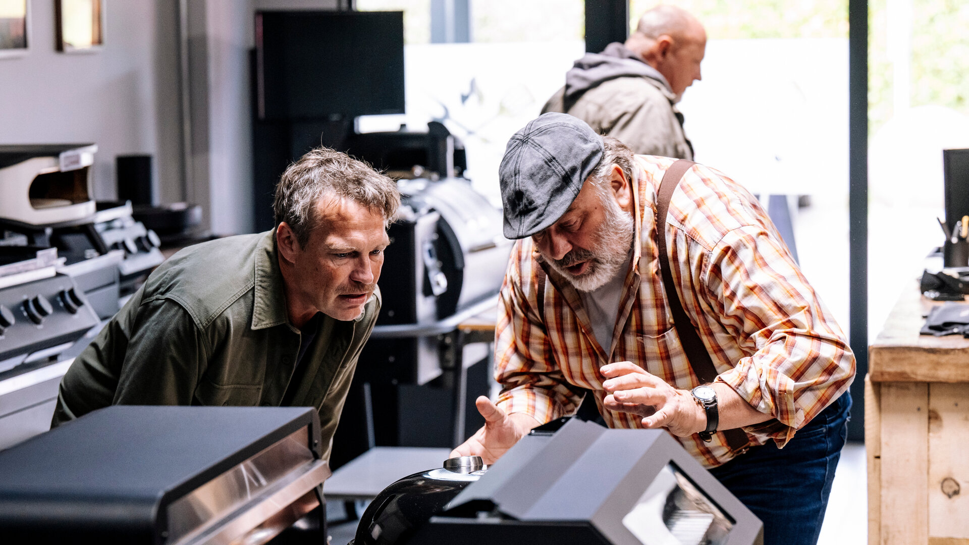 "SOKO Köln - Doppelt gestorben": Matti Wagner (Pierre Besson) und Stefan Ständler (Waldemar Kobus) stehen dicht beieinander in einem Geschäft. Beide blicken konzentriert auf einen Grill, der vor ihnen steht. Stefan Ständler gestikuliert beim Reden mit den Händen. Die Umgebung ist funktional eingerichtet, mit Regalen und Geräten im Hintergrund. Im Hintergrund läuft ein Mann (Komparse) durch den Laden.