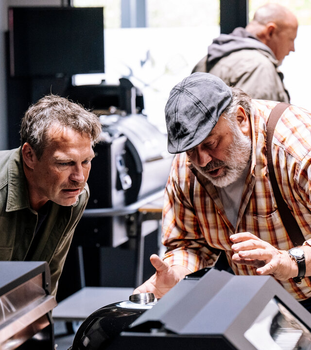 "SOKO Köln - Doppelt gestorben": Matti Wagner (Pierre Besson) und Stefan Ständler (Waldemar Kobus) stehen dicht beieinander in einem Geschäft. Beide blicken konzentriert auf einen Grill, der vor ihnen steht. Stefan Ständler gestikuliert beim Reden mit den Händen. Die Umgebung ist funktional eingerichtet, mit Regalen und Geräten im Hintergrund. Im Hintergrund läuft ein Mann (Komparse) durch den Laden.