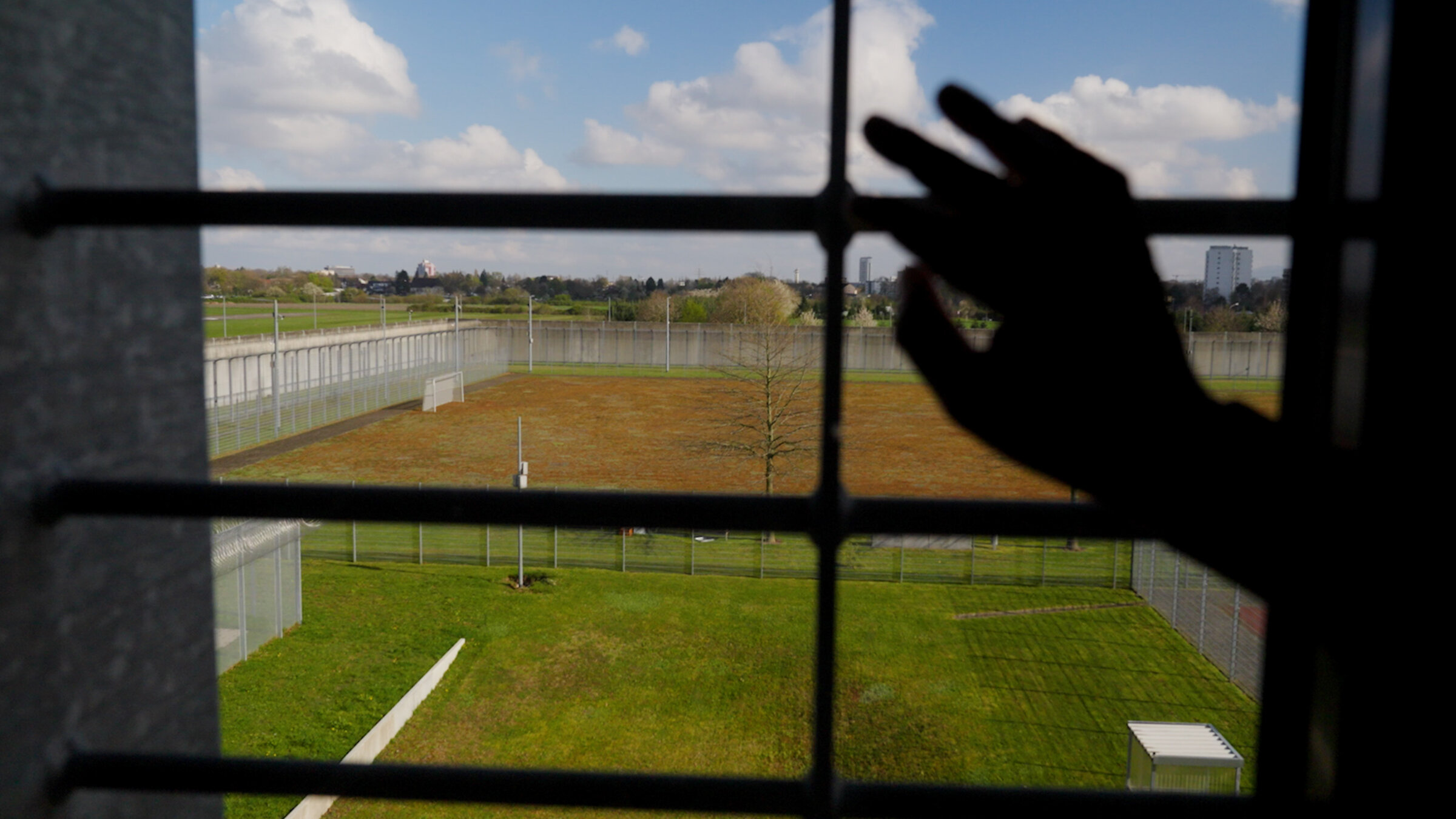 "Im Gefängnis: Das Prinzip Hoffnung": Eine Hand fasst an ein vergittertes Fenster. Hinter dem Fenster liegt ein großer Platz mit Rasen, der von einem hohen Zaun umfasst ist.