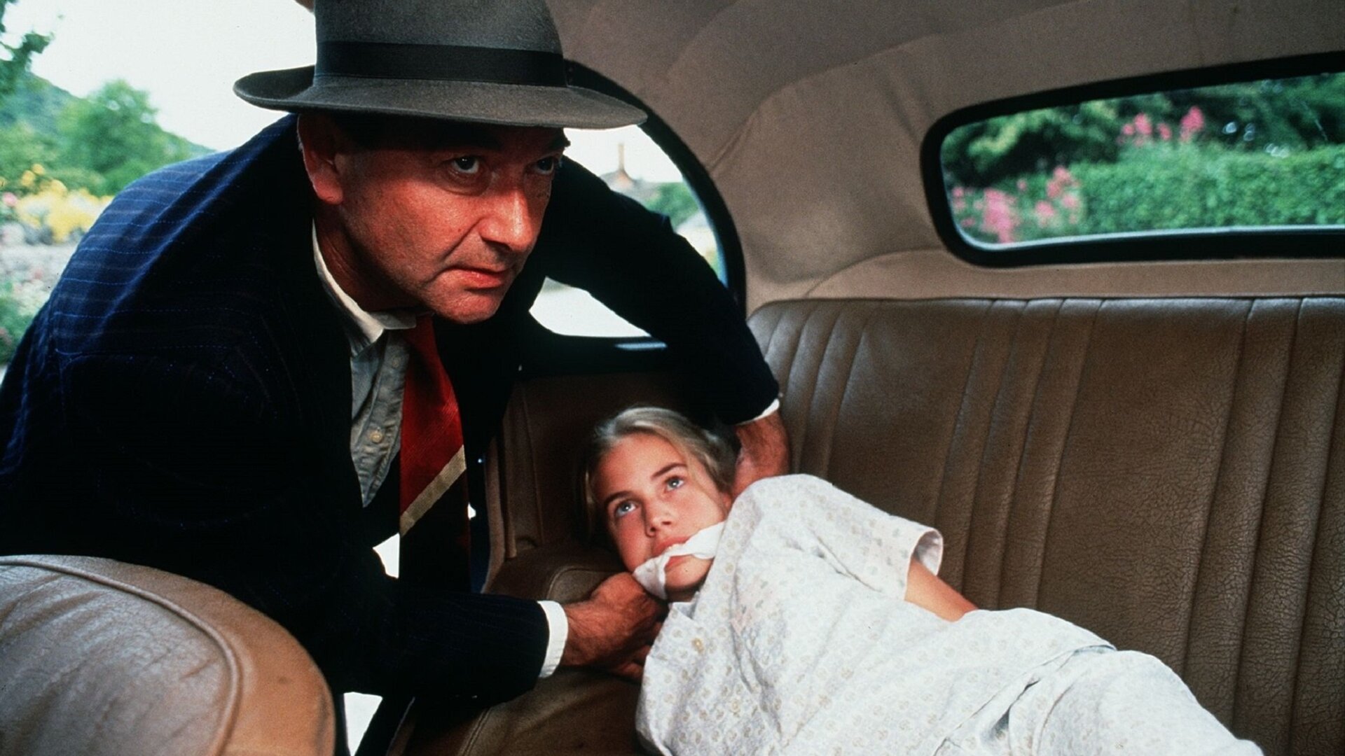 "Fünf Freunde auf geheimnisvollen Spuren": Mr. Stock (Peter Jonfield) lehnt sich in ein Auto. Dabei trägt er einen Anzug und einen Hut und hat seine Hände um ein Tuch, dass er Jennifer Armstrong (Philippa Woodward) um den Mund gebunden hat. Jennifer  Armstrong (Philippa Woodward) liegt in einem Schlafanzug auf der Rückbank des Autos.