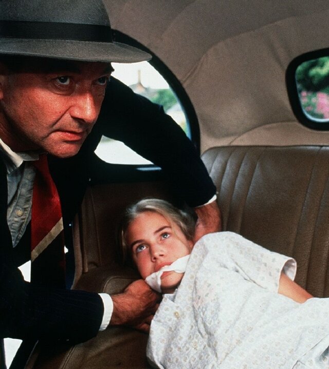 "Fünf Freunde auf geheimnisvollen Spuren": Mr. Stock (Peter Jonfield) lehnt sich in ein Auto. Dabei trägt er einen Anzug und einen Hut und hat seine Hände um ein Tuch, dass er Jennifer Armstrong (Philippa Woodward) um den Mund gebunden hat. Jennifer  Armstrong (Philippa Woodward) liegt in einem Schlafanzug auf der Rückbank des Autos.