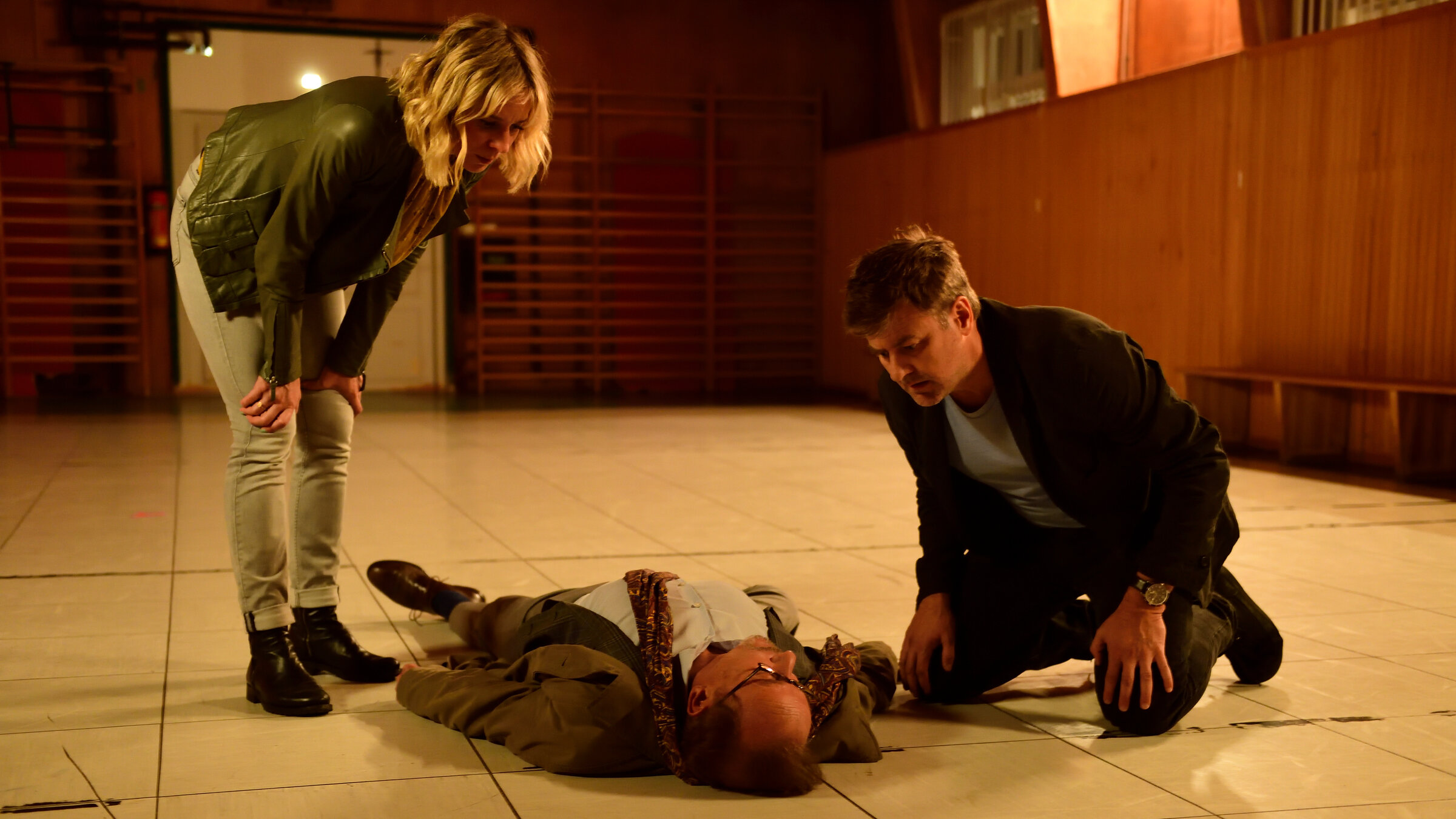 "München Mord - Die indische Methode": In einer Turnhalle: Ludwig Schaller (Alexander Held) liegt reglos auf dem Boden. Angela Flierl (Bernadette Heerwagen) ist über ihn gebeugt und Harald Neuhauser (Marcus Mittermeier) kniet neben ihm. Beide schauen ihn von oben an.