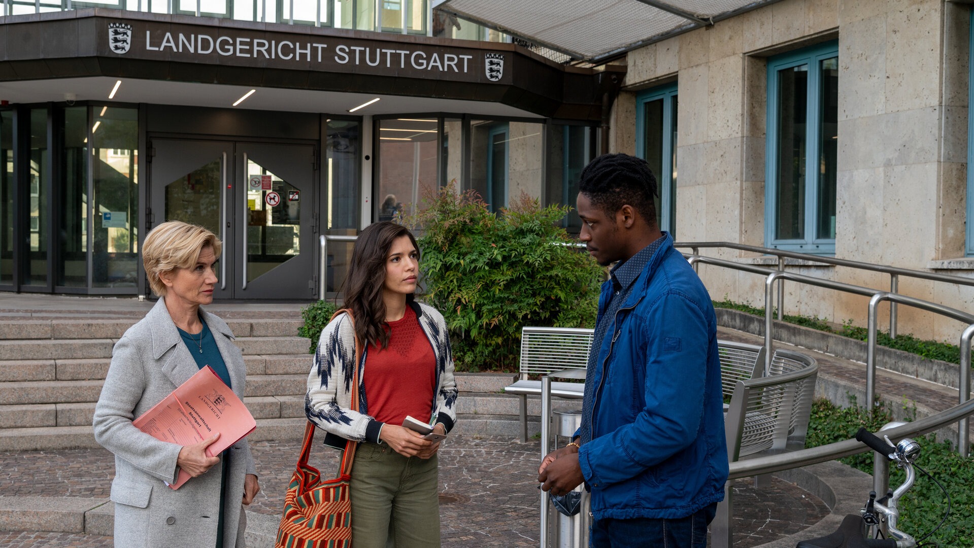 "SOKO Stuttgart - Straflust": Martina Seiffert (Astrid M. Fünderich) und Lea Gomez (Jasmin Gassmann) stehen vor dem Landgericht Stuttgart. Ihnen gegenüber steht Essam Keita (Ben Andrews Rumler). Martina hält eine Akte, Lea ihr Notizbuch in den Händen. Die beiden blicken Essam streng an, dieser wirkt eingeschüchtert.