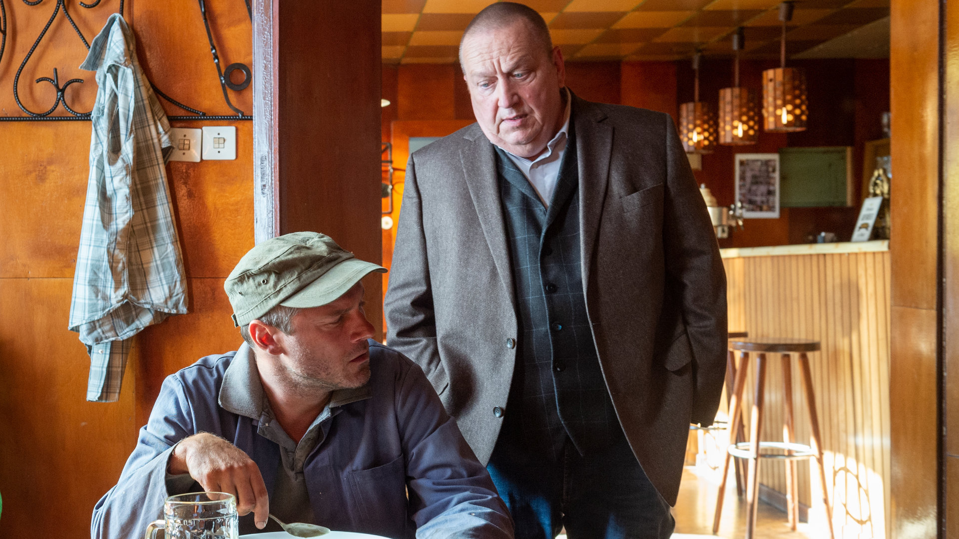 "SOKO Wismar – Gemeiner Stechapfel": Lukas Hentsch (Jörg Malchow) sitzt am Tisch in einer Kneipe. Vor sich hat er einen gefüllten Teller mit Suppe. Revierchef Reuter (Udo Kroschwald) steht hinter ihm und wirft ihm einen auffordernden Blick zu. Lukas Hentsch wirkt kritisch gegenüber Reuter.