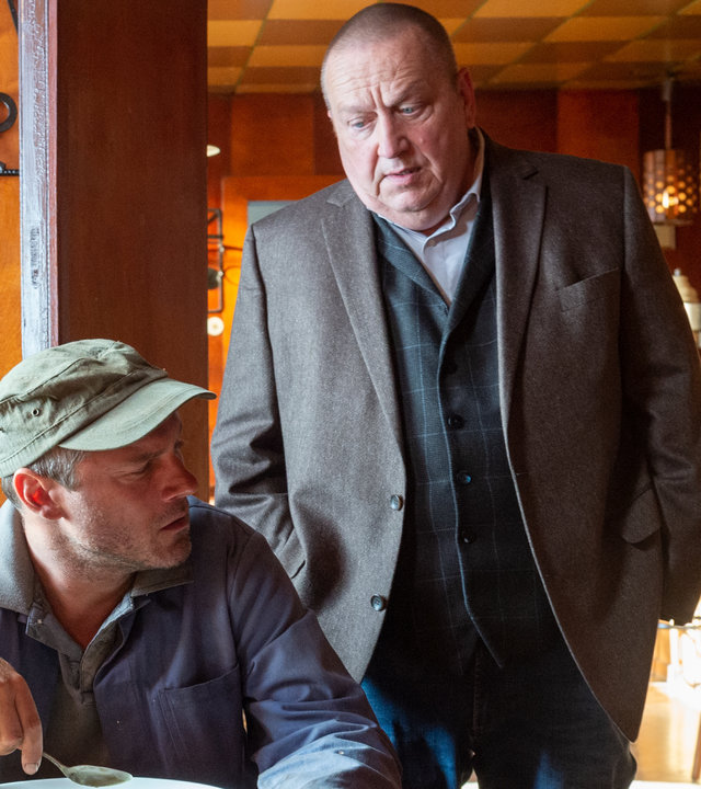 "SOKO Wismar – Gemeiner Stechapfel": Lukas Hentsch (Jörg Malchow) sitzt am Tisch in einer Kneipe. Vor sich hat er einen gefüllten Teller mit Suppe. Revierchef Reuter (Udo Kroschwald) steht hinter ihm und wirft ihm einen auffordernden Blick zu. Lukas Hentsch wirkt kritisch gegenüber Reuter.