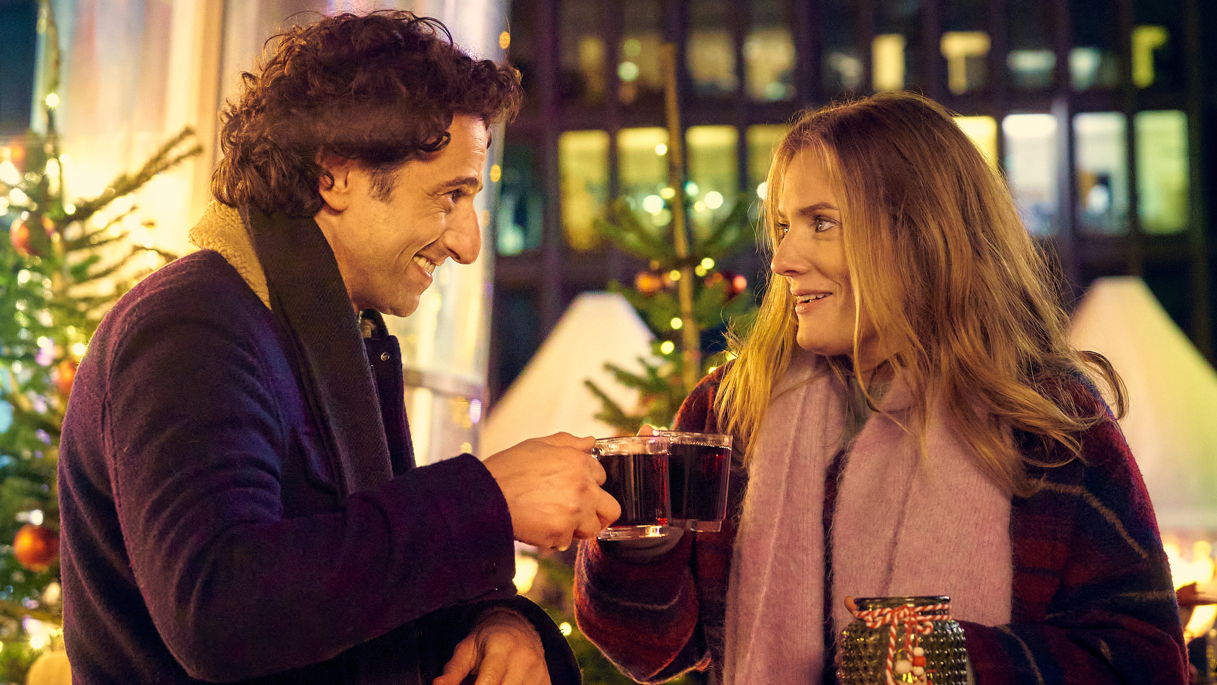 "Zwei Weihnachtsmänner sind einer zu viel": Glücklich blicken sich Karo (Marie Burchard) und Lasse (Serkan Kaya) in die Augen. Sie stehen an einem roten Stehtisch mit Weihnachtsdeko und stoßen mit Glühweingläsern an. Im Hintergrund stehen zwei leuchtende Weihnachtsbäume.