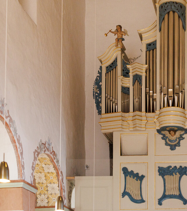 "Evangelischer Gottesdienst - Wenn dein Kind dich morgen fragt...": Im Innenraum der evangelischen Kirche St. Severus in Gemünden steht die Orgel: Sie ist weiß mit goldenen Umrandungen und blauen Intarsien versehen. Auf beiden Seiten der Orgel sitzen kleine trompetende Engel. An der Seite sind die mit Blumenranken bemalten Arkadenbögen der Kirche zu sehen.