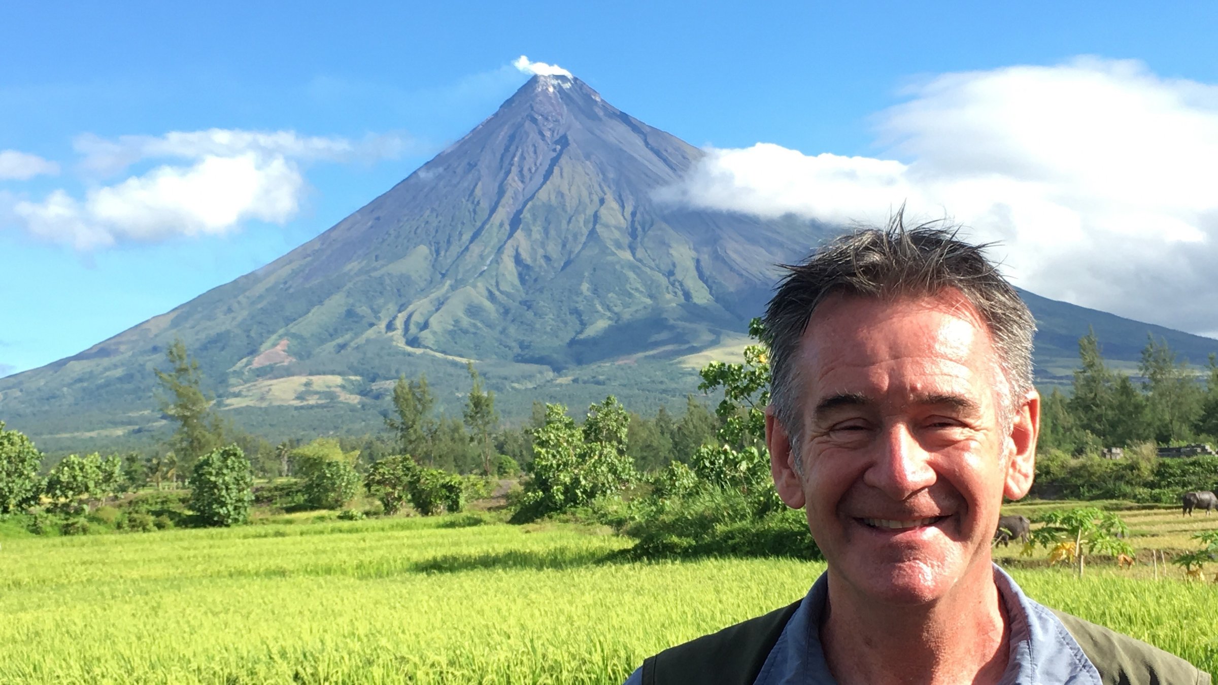 "Die wilden Philippinen: Der vulkanische Norden": Nahaufnahme von Nigel Marven. Im Hintergrund befindet sich der Vulkan Mayon, davor grüne Felder, Bäume und Büsche.