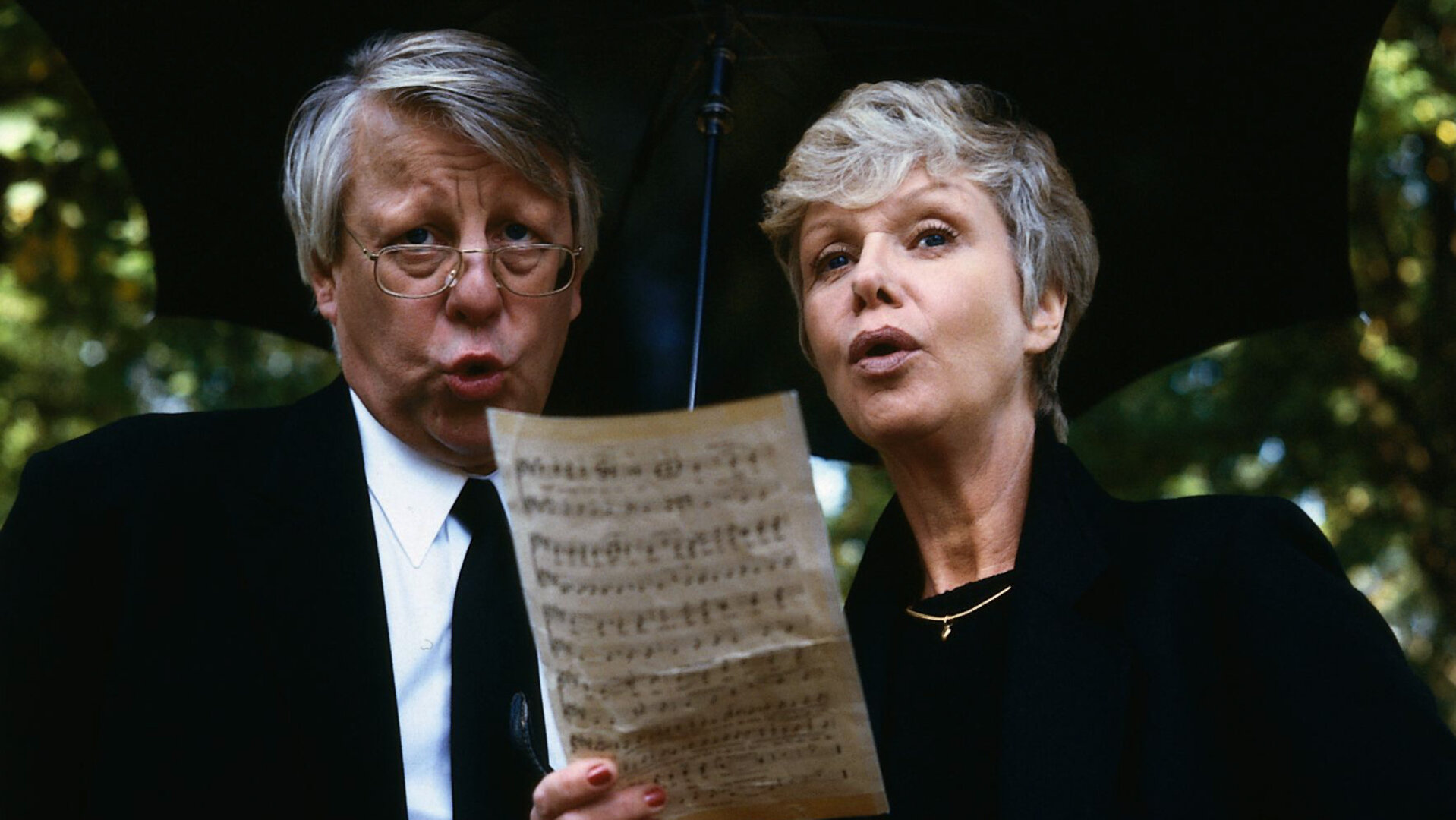 "Die Wicherts von nebenan: Berlin, Herzogstr. 36": Eberhard (Stefan Orlac) und Hannelore Wichert (Maria Sebaldt) stehen unter einem Regenschirm und halten ein Notenblatt in der Hand. Beide tragen schwarze Kleidung und singen.