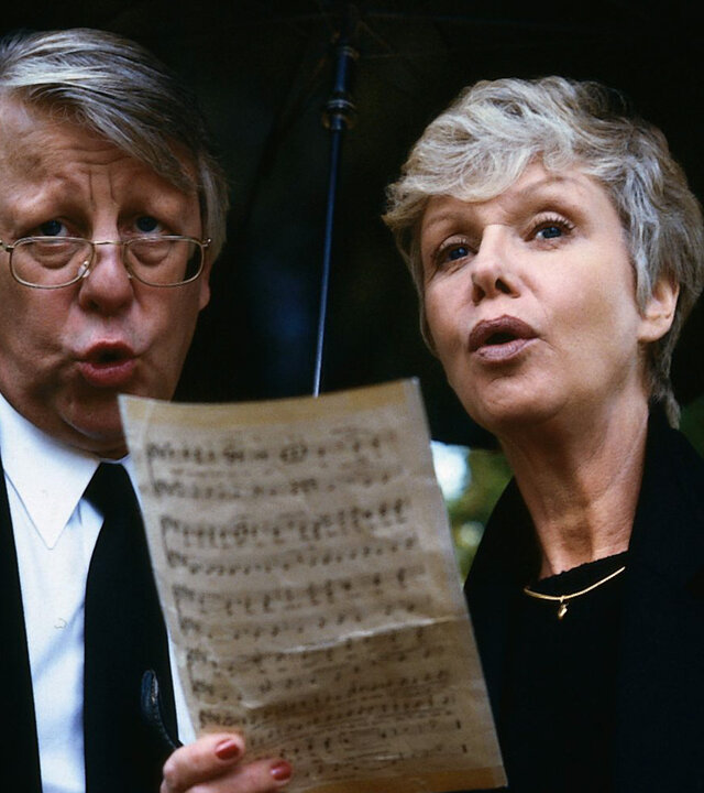 "Die Wicherts von nebenan: Berlin, Herzogstr. 36": Eberhard (Stefan Orlac) und Hannelore Wichert (Maria Sebaldt) stehen unter einem Regenschirm und halten ein Notenblatt in der Hand. Beide tragen schwarze Kleidung und singen.
