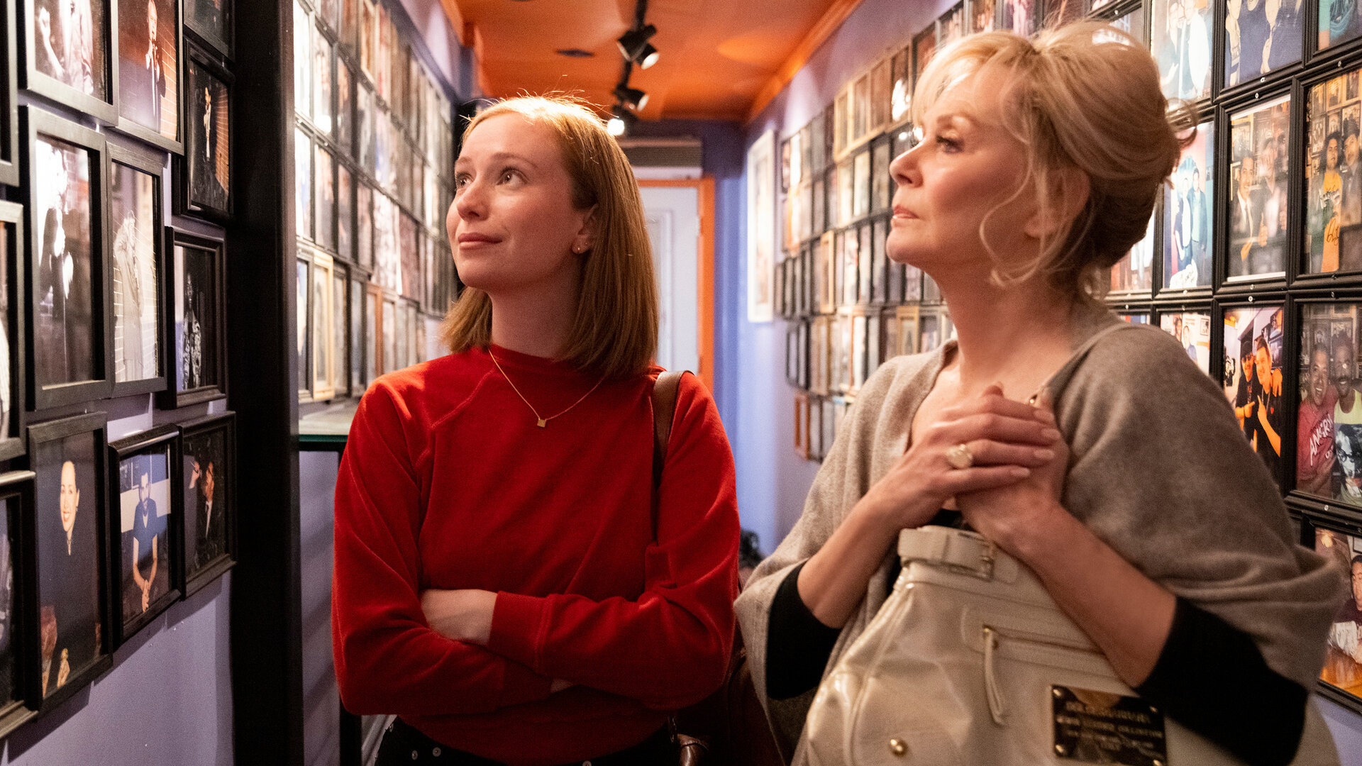 "Hacks - 1,69 Millionen": Ava ((Hannah Einbinder) und Deborah (Jean Smart) stehen in einem schmalen Flur, dessen Wände dicht mit gerahmten Fotografien bedeckt sind. Während Ava die Fotowand leicht lächelnd betrachtet, wirft Deborah einen nachdenklichen Blick darauf.