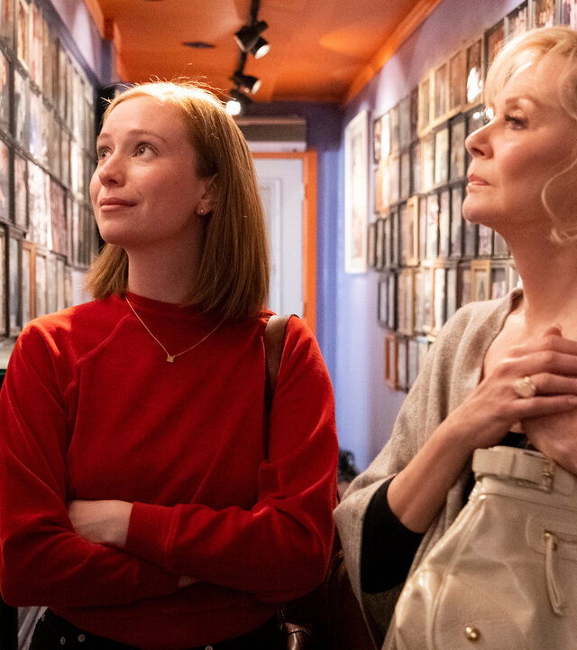 "Hacks - 1,69 Millionen": Ava ((Hannah Einbinder) und Deborah (Jean Smart) stehen in einem schmalen Flur, dessen Wände dicht mit gerahmten Fotografien bedeckt sind. Während Ava die Fotowand leicht lächelnd betrachtet, wirft Deborah einen nachdenklichen Blick darauf.