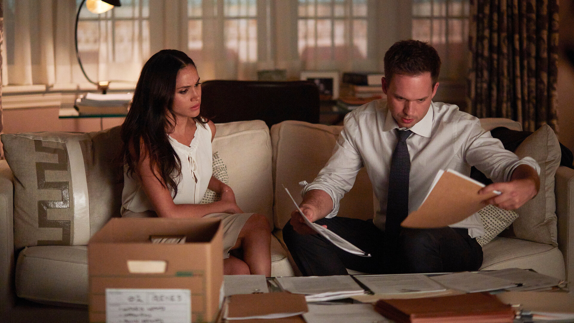"Suits - Karten auf den Tisch": Rachel (Meghan Markle) und Mike (Patrick J. Adams) sitzen nebeneinander auf einem Sofa und schauen Dokumente an, die Mike in beiden Händen hält. Beide haben einen besorgten Gesichtsausdruck. Vor ihnen auf dem Tisch eine offene Kiste und ausgebreitete Dokumente.
