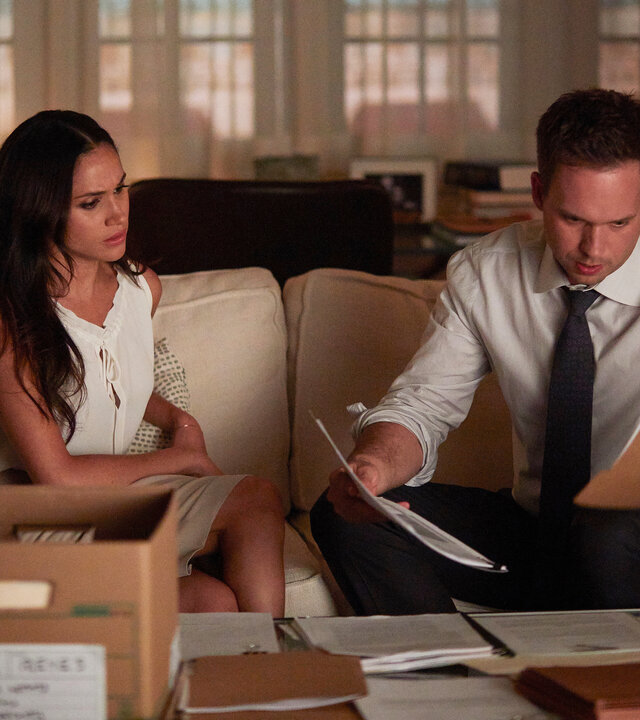 "Suits - Karten auf den Tisch": Rachel (Meghan Markle) und Mike (Patrick J. Adams) sitzen nebeneinander auf einem Sofa und schauen Dokumente an, die Mike in beiden Händen hält. Beide haben einen besorgten Gesichtsausdruck. Vor ihnen auf dem Tisch eine offene Kiste und ausgebreitete Dokumente.