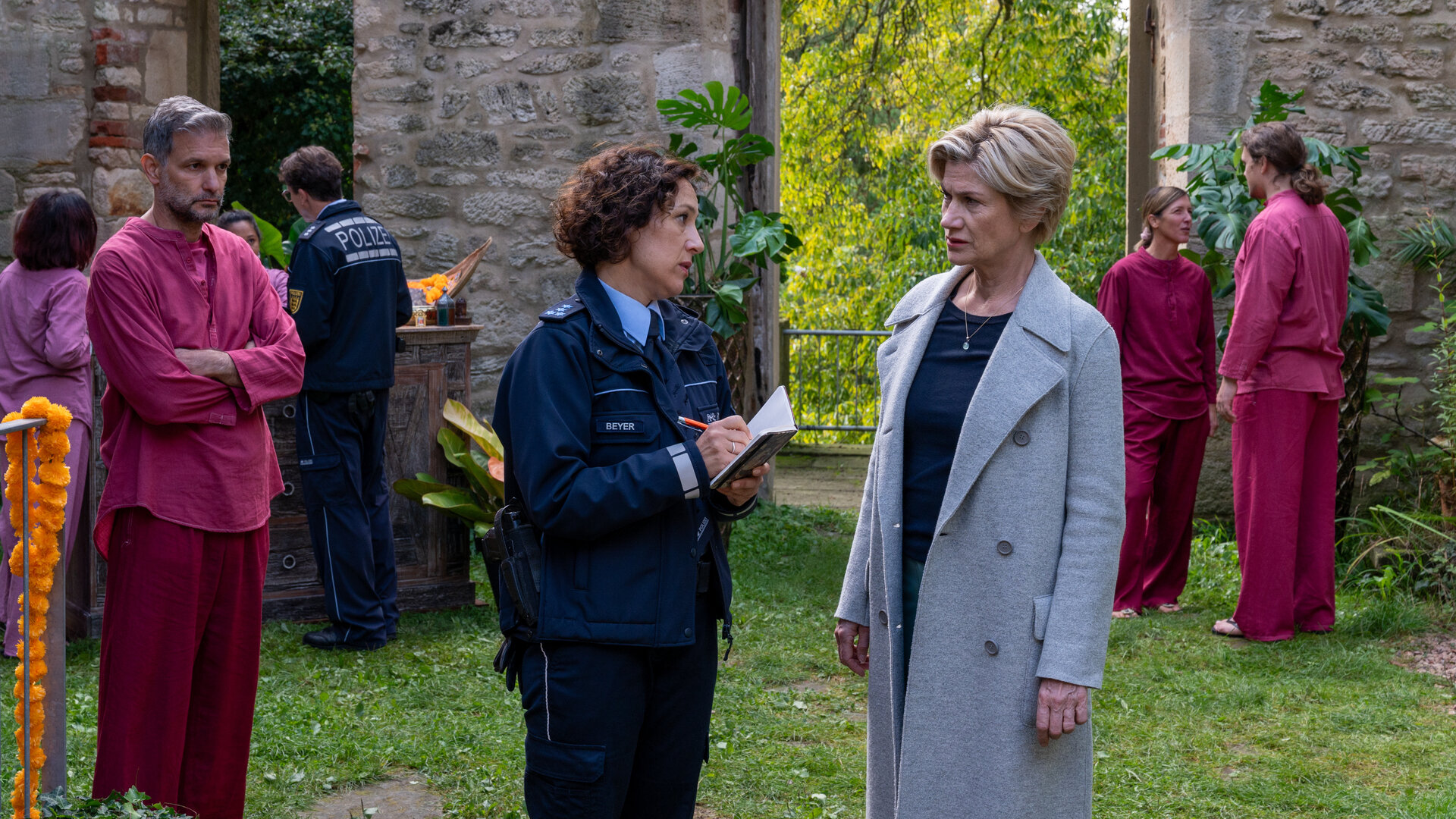 "SOKO Stuttgart - Der Kuss des Frosches": Die erste Kriminalkommissaren Martina Seiffert (Astrid M. Fünderich) und Streifenpolizistin Sibylle Beyer (Bärbel Stolz) stehen im begrünten Innenhof eines Gebäudes, das wie eine Burgruine aussieht. Die beiden Frauen schauen sich ernst an. Im Hintergrund stehen mehrere Personen (Komparsen), die in weinrote Leinenhosen und Oberteile gekleidet sind. Einer von ihnen, Veit Kerstens (Luca A. Zamperoni), steht mit verschränkten Armen ganz in der Nähe von Martina und Bille und scheint deren Gespräch zu lauschen.