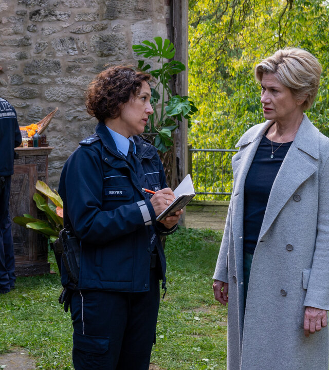 "SOKO Stuttgart - Der Kuss des Frosches": Die erste Kriminalkommissaren Martina Seiffert (Astrid M. Fünderich) und Streifenpolizistin Sibylle Beyer (Bärbel Stolz) stehen im begrünten Innenhof eines Gebäudes, das wie eine Burgruine aussieht. Die beiden Frauen schauen sich ernst an. Im Hintergrund stehen mehrere Personen (Komparsen), die in weinrote Leinenhosen und Oberteile gekleidet sind. Einer von ihnen, Veit Kerstens (Luca A. Zamperoni), steht mit verschränkten Armen ganz in der Nähe von Martina und Bille und scheint deren Gespräch zu lauschen.