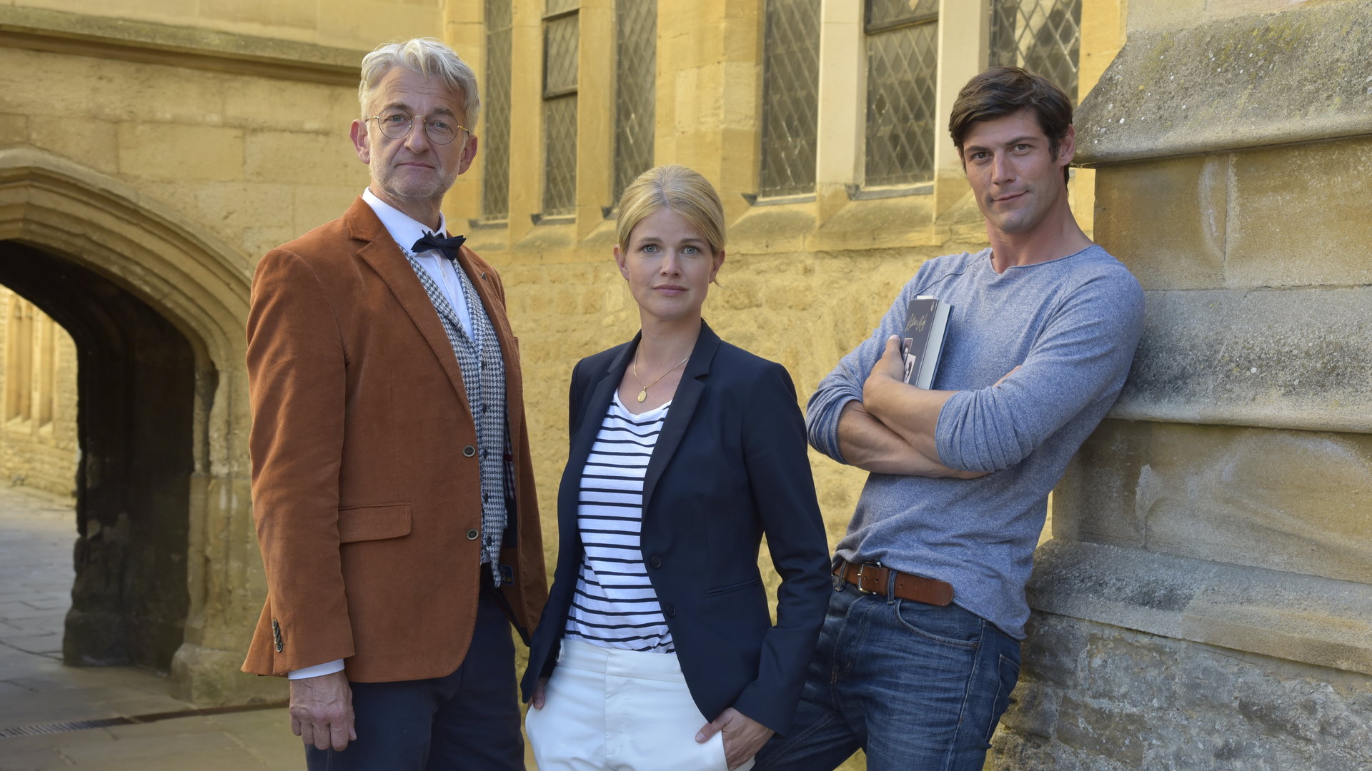 "Ein Sommer in Oxford": Nina (Mira Bartuschek) steht zwischen den beiden Männern Daniel Miller (Michael Raphael Klein) und Prof. Thomas Rothwell (Dominic Raacke) vor einer Kirche. Thomas blickt direkt in die Kamera, Nina hat die Hände lässig in den Hosentaschen und Daniel hat die Arme vorne verschränkt.
