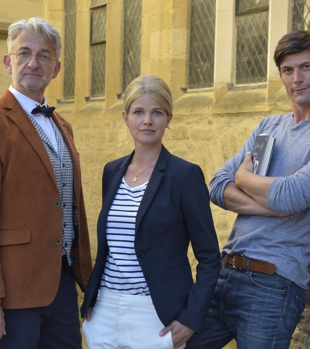 "Ein Sommer in Oxford": Nina (Mira Bartuschek) steht zwischen den beiden Männern Daniel Miller (Michael Raphael Klein) und Prof. Thomas Rothwell (Dominic Raacke) vor einer Kirche. Thomas blickt direkt in die Kamera, Nina hat die Hände lässig in den Hosentaschen und Daniel hat die Arme vorne verschränkt.