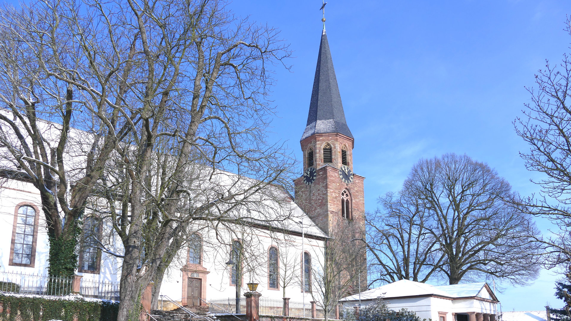 "Katholischer Gottesdienst - Die Liebe weitertragen": Außenansicht der Kirche Mariä Himmelfahrt in Herxheim im Winter, es liegt bei Schnee.