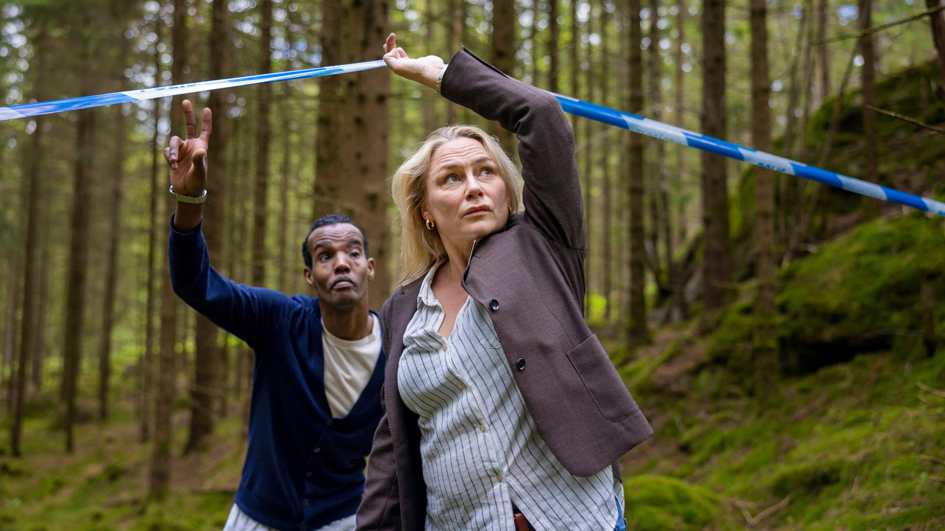 "Detective No. 24: Hase und Igel": Ibraahin (Nasir Farah Dhagole) und Tilda (Malin Levanon) sind im Wald. Tilda hebt das Absperrband der Polizei an und geht darunter durch. Ibraahin folgt ihr.