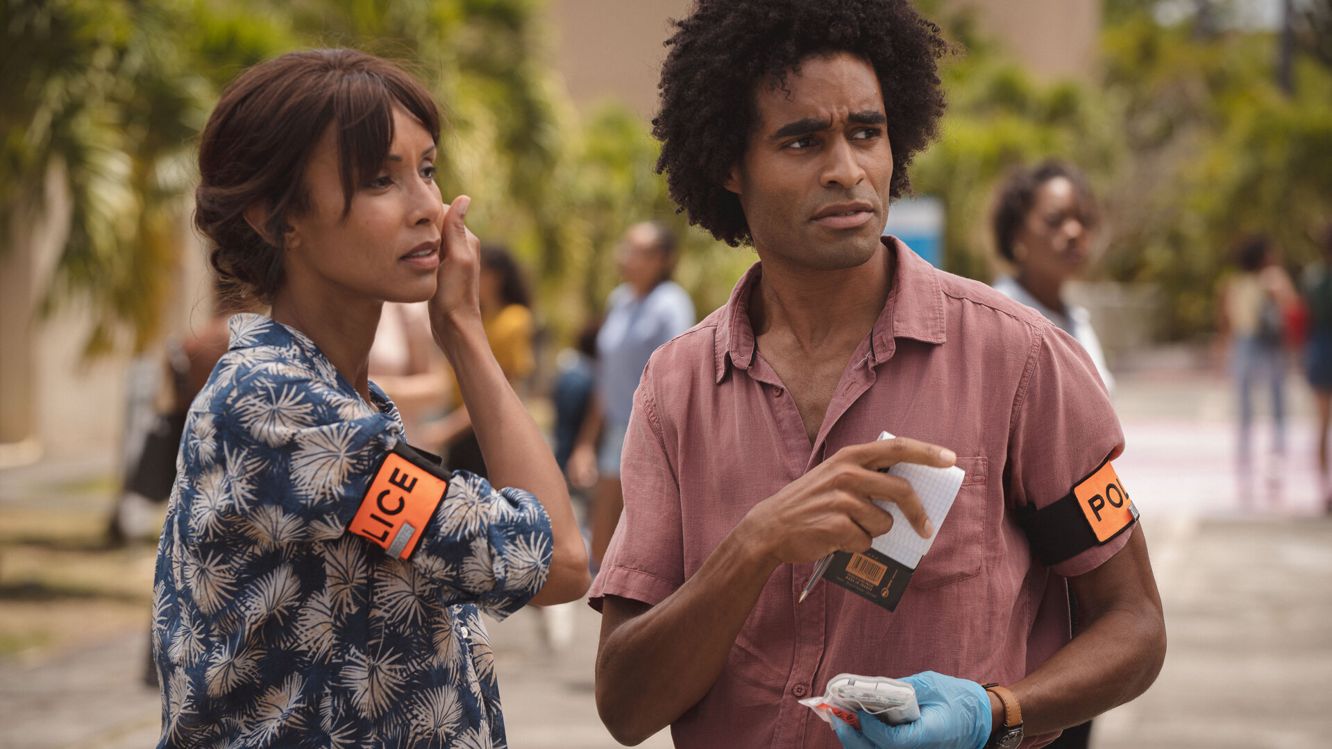 "Deadly Tropics - Das schnelle Geld": Mélissa (Sonia Rolland) und Aurélien (Julien Béramis) stehen in einer städtischen Umgebung und schauen aufmerksam zur Seite. Aurélien ist mit einem Untersuchungshandschuh sowie einem Notizblock und einem Stift ausgestattet.