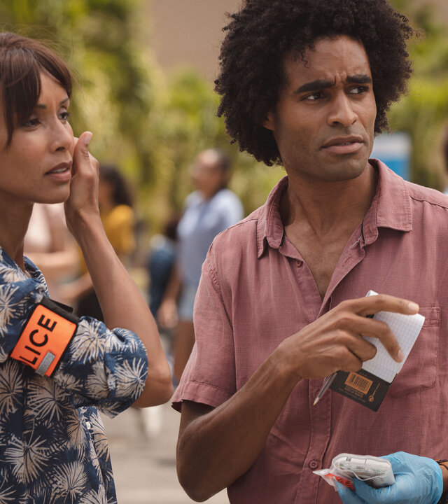 "Deadly Tropics - Das schnelle Geld": Mélissa (Sonia Rolland) und Aurélien (Julien Béramis) stehen in einer städtischen Umgebung und schauen aufmerksam zur Seite. Aurélien ist mit einem Untersuchungshandschuh sowie einem Notizblock und einem Stift ausgestattet.