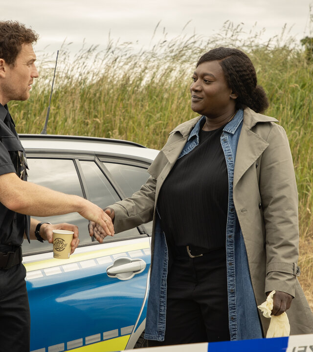 "Die Polizistin und die Sprache des Todes": Pieper Olsen (Artjom Gilz) und Gloria Acheampong (Thelma Buabeng) stehen neben einem Polizeiwagen und geben sich die Hand.