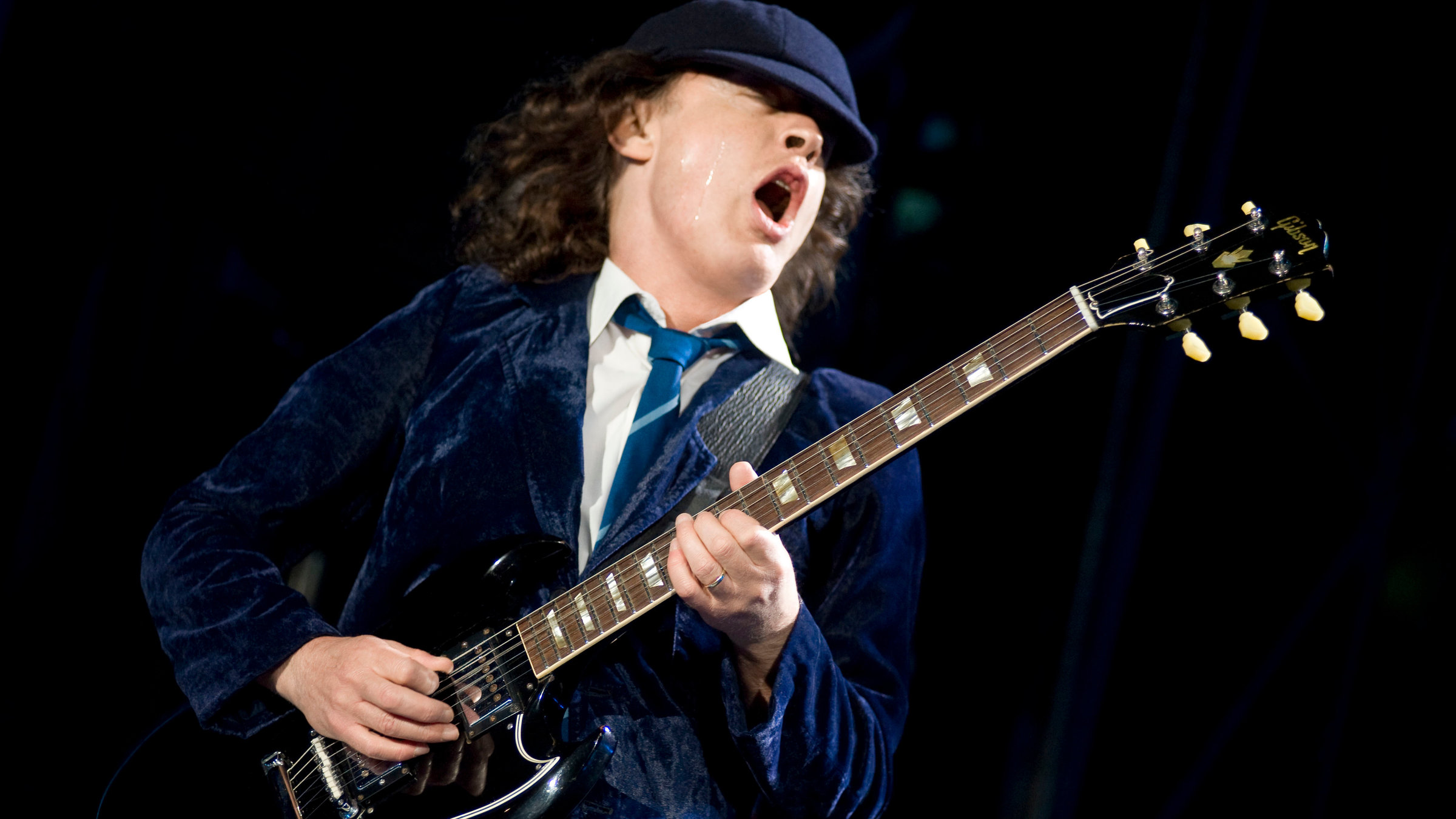 "Pop Around the Clock – Summer Edition: AC/DC: Live at Donington": Ein Mann mit dunkelblauem Samtsakko, Krawatte und weißem Hemd spielt auf einer E-Gitarre und singt mit geschlossenen Augen und weit geöffnetem Mund.
