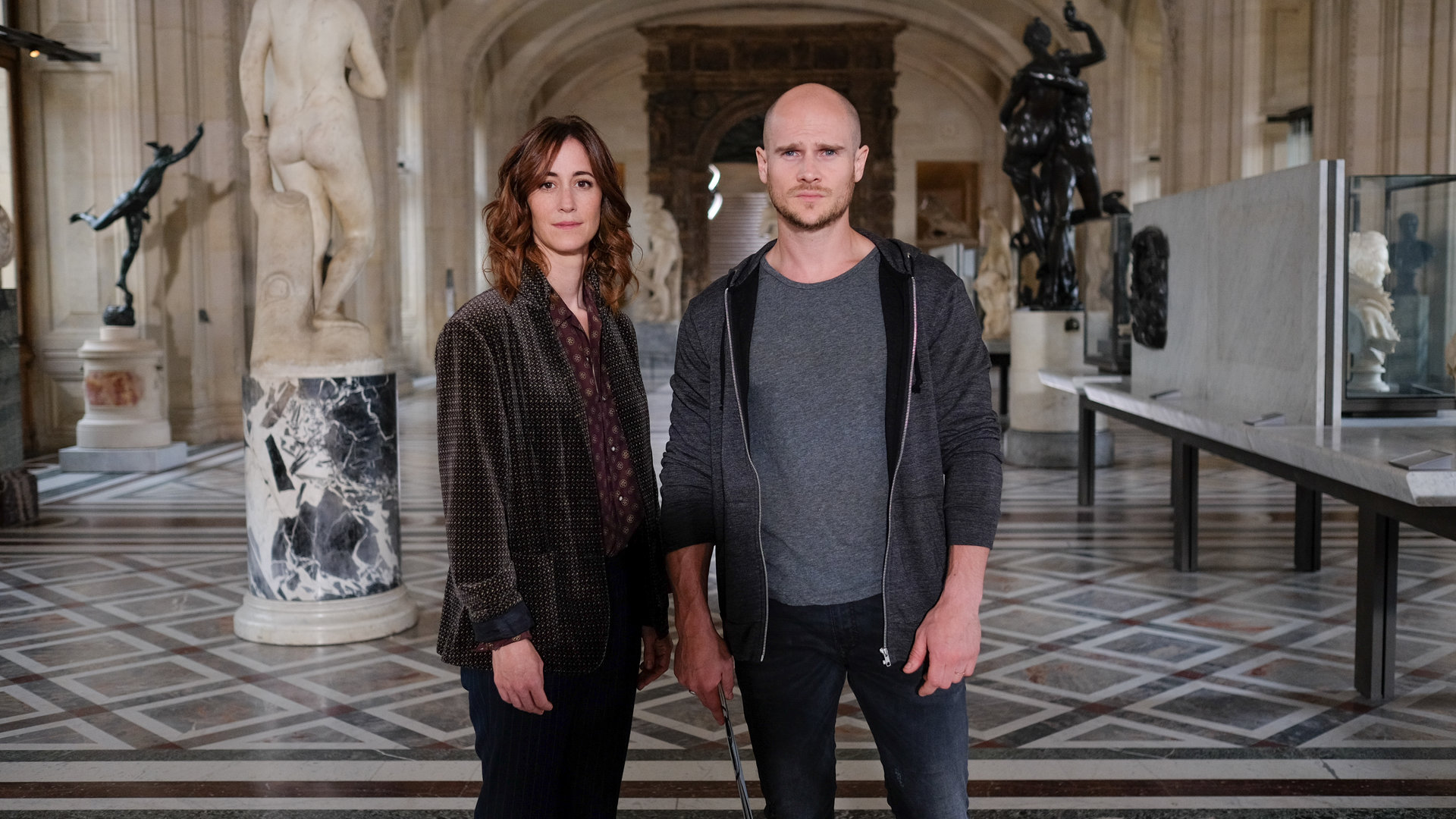 "Art of Crime - Der Da Vinci Code (1)": Florence Chassagne (Éléonore Gosset) und Antoine Verlay (Nicolas Gob) stehen in einem großen Museumsraum mit antiken Statuen und Schaukästen und schauen in die Kamera.