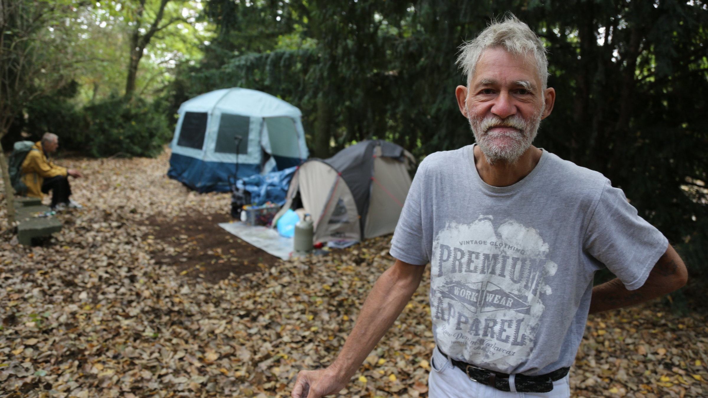 "ZDF.reportage: Armes reiches Deutschland - Verteilungskampf": Der Obdachlose Peter, ein Mann mit grauen Haaren und Vollbart, trägt ein graues T-Shirt mit Aufdruck. Er steht im Vordergrund und schaut in die Kamera. Hinten stehen zwei Zelte unter Nadelbäumen und ein Mann mit gelber Jacke sitzt auf einer Bank. Laub liegt auf dem Boden.