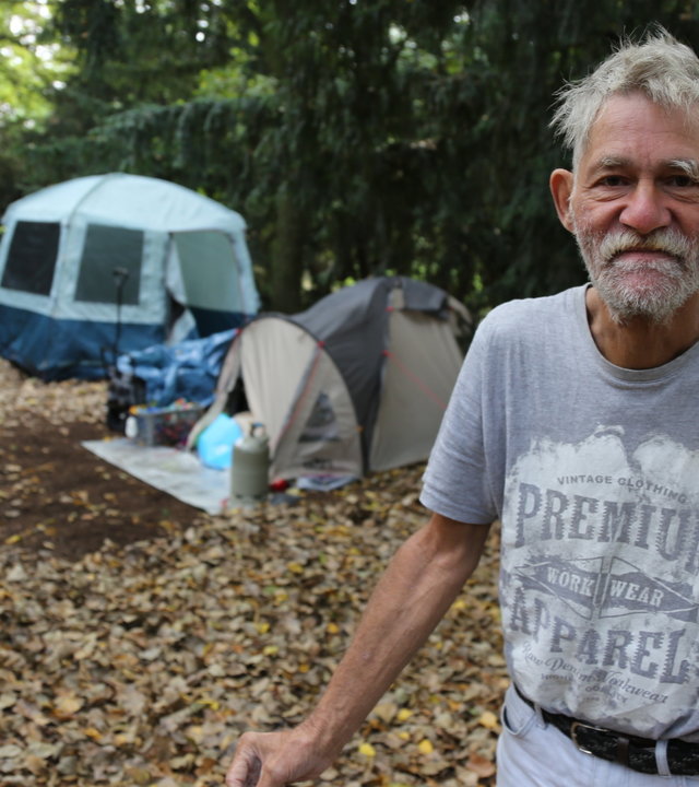 "ZDF.reportage: Armes reiches Deutschland - Verteilungskampf": Der Obdachlose Peter, ein Mann mit grauen Haaren und Vollbart, trägt ein graues T-Shirt mit Aufdruck. Er steht im Vordergrund und schaut in die Kamera. Hinten stehen zwei Zelte unter Nadelbäumen und ein Mann mit gelber Jacke sitzt auf einer Bank. Laub liegt auf dem Boden.