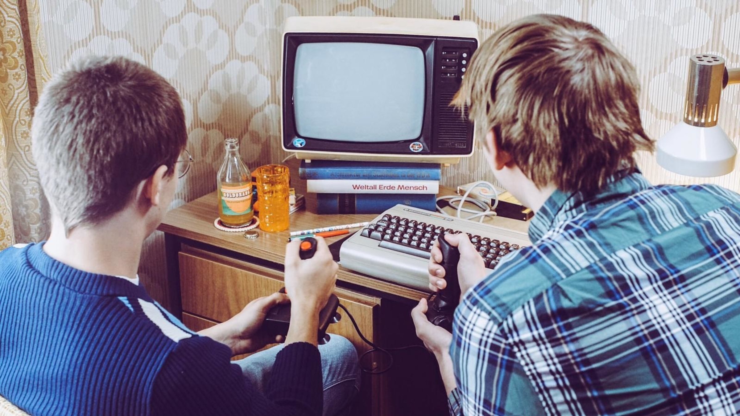 "Auferstanden aus Platinen": Zwei Jugendliche sitzen mit ihren Joysticks vor einer Computertastatur und einem Fernseher aus den 80er Jahren. Möbel und Tapete sind aus den 80er Jahren.