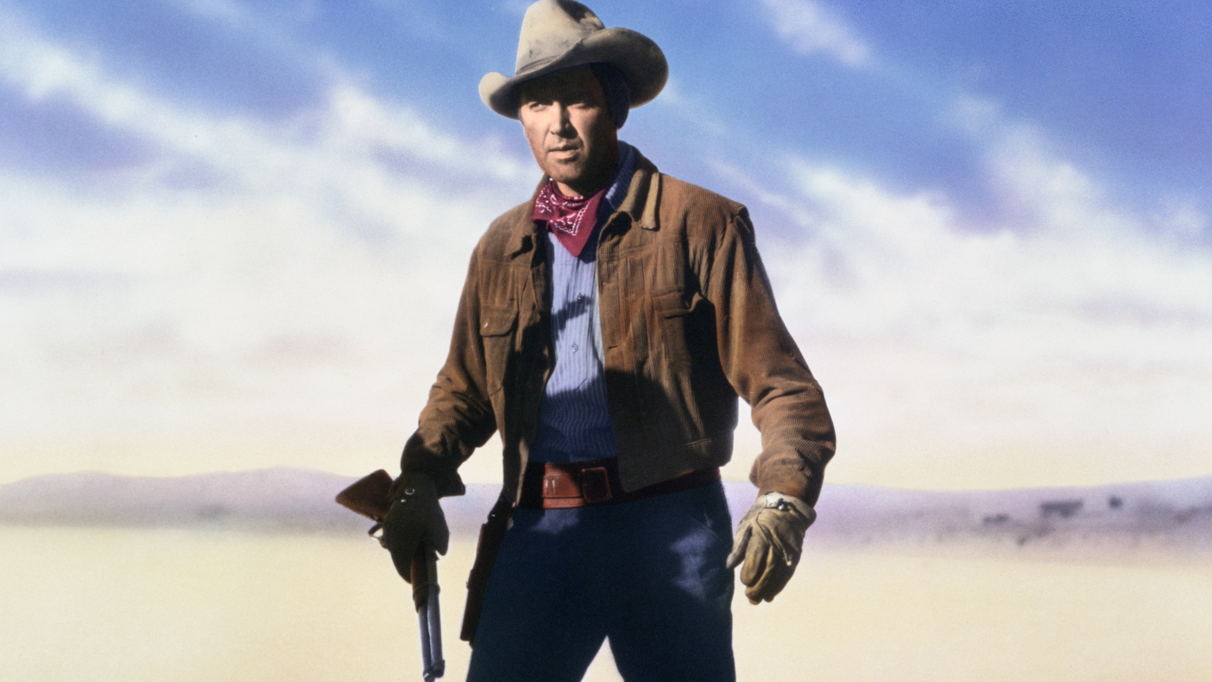 "Der Mann aus Laramie“: Vor einer weiten, ebenen Landschaft steht Will Lockhart (James Stewart) in typischer Cowboy-Kluft und mit entschlossenem Blick. In seiner rechten Hand hält er locker ein Gewehr, dessen Lauf nach unten zeigt.