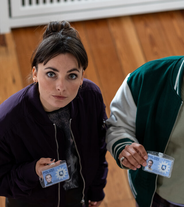 "SOKO Hamburg - Schlechte Karten": Blick von schräg oben: Franzi Berger (Paula Schramm) und Luiz Vieira dos Santos (Thiago Braga de Oliveira) stehen nebeneinander auf einem Parkettboden und schauen nach oben. Sie halten mit ernstem Blick ihre Dienstausweise sichtbar vor den Körper.