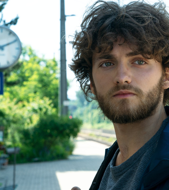 Am Ende - Die Macht der Kränkung - Leon": Leon (Léon Orlandianyi) steht vor der Kamera und blickt in die Ferne. Im Hintergrund ist ein Bahnhof zu sehen und eine große Uhr, die an der Hausfassade befestigt ist.