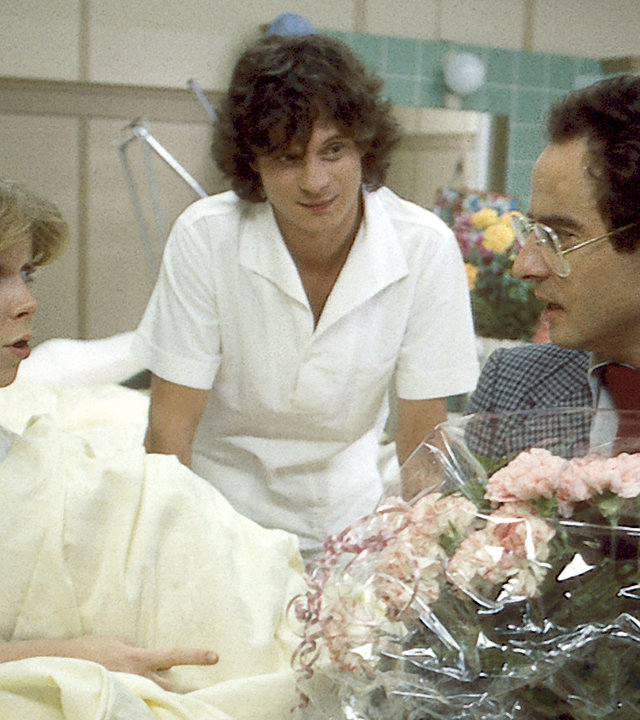 "Die Schwarzwaldklinik - Vaterschaft": Stefan (Volker Eckstein) sitzt mit einem großen Blumenstrauß bei Franziska (Marion Kracht) am Krankenbett. Sie wirkt aufgeregt. Ein Pfleger steht außerdem am Bett.