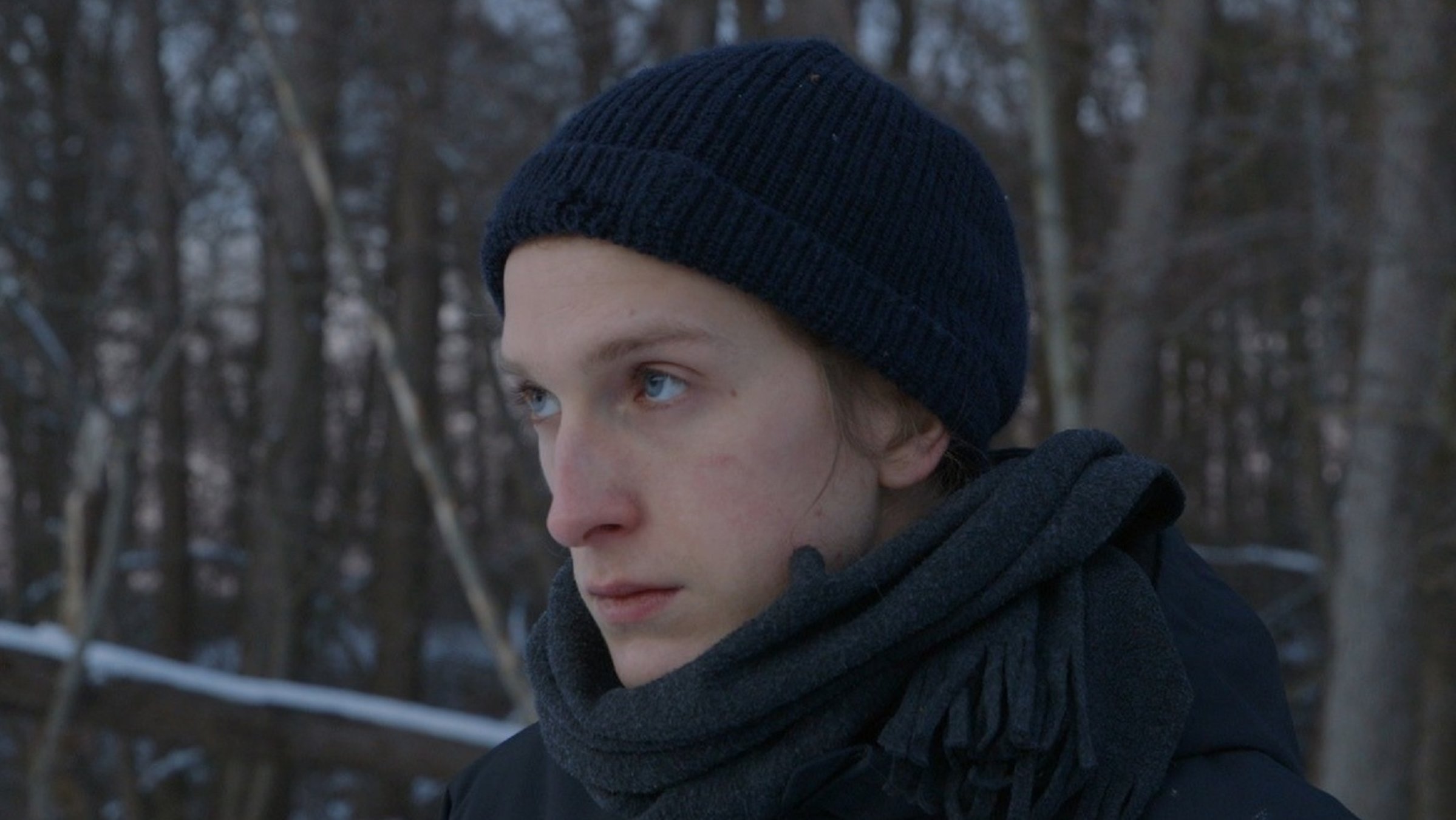 "Ab 18! - Karls Freiheit: Großaufnahme des Kopfes eines bartlosen jungen Mannes, der eine blaue Wollmütze und einen blauen Schal trägt und nachdenklich, in die Ferne schaut. Er steht in einer winterlichen Landschaft.