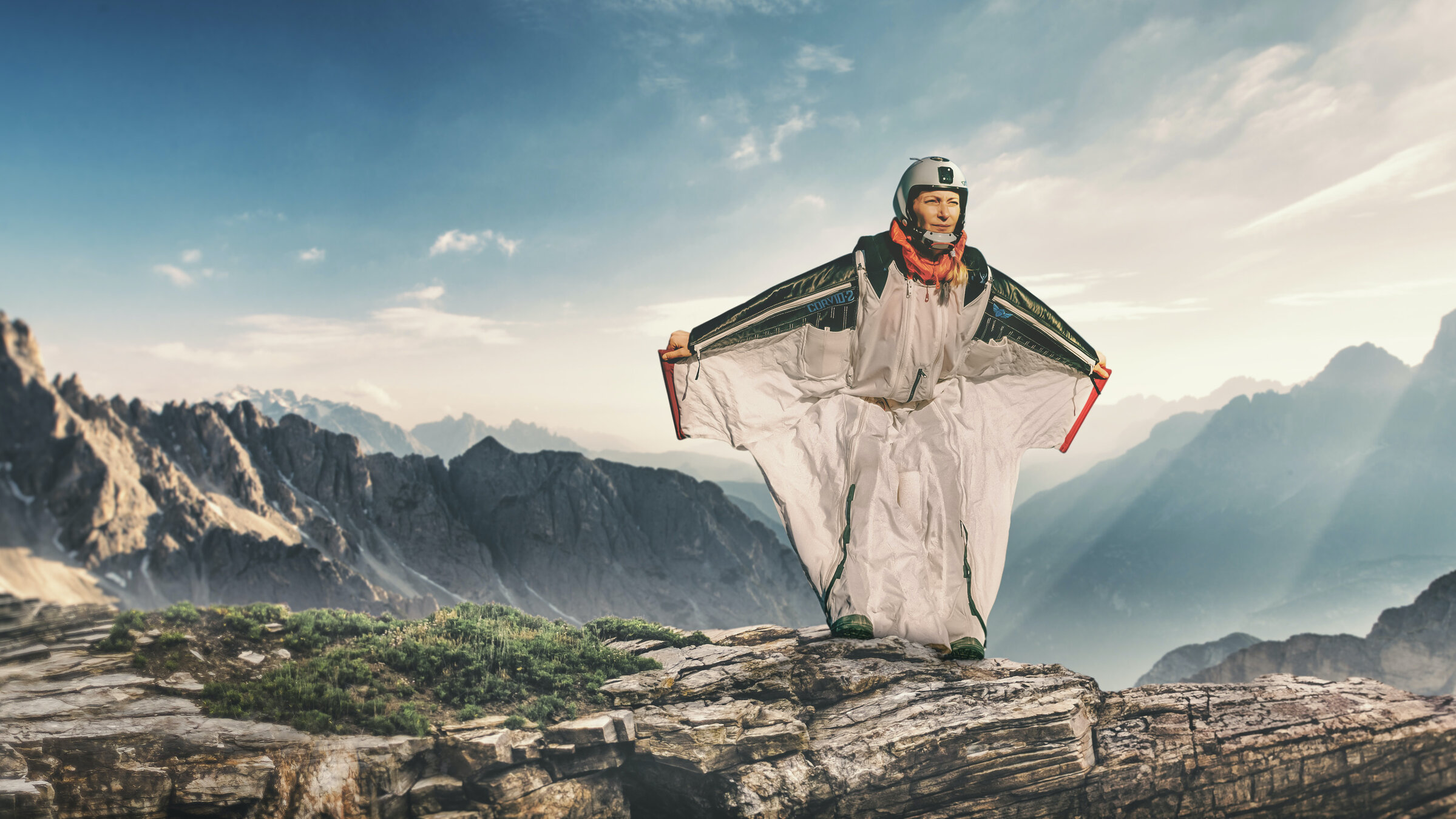 "Adrenalin pur. Wenn Frauen ans Limit gehen": Katja steht in ihrem Wingsuit (Flügelanzug) oben auf einem Berg. Hinter ihr liegen weitere hohen Berge im Sonnenschein.