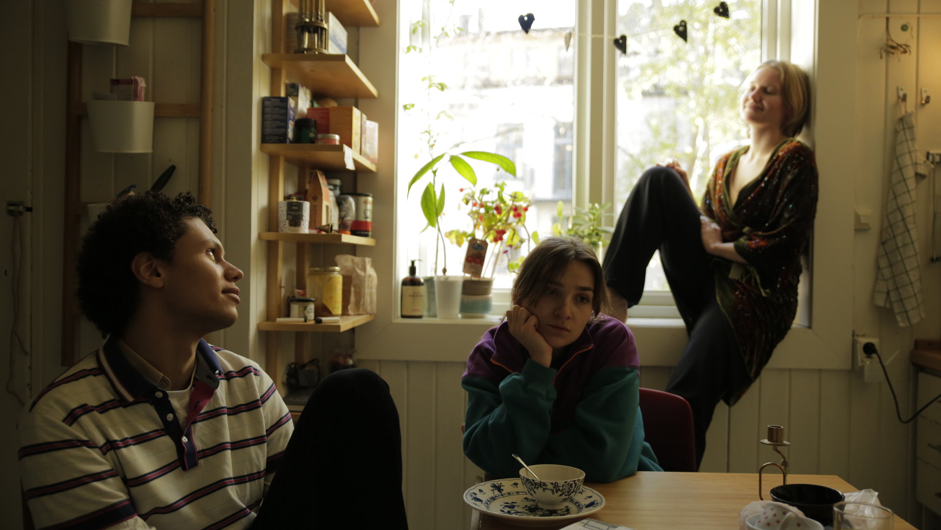 "About Saturday - Red mit mir": Lotte (Hanna Heider Hov), Klara (Darin Hagi) und Mo (Kayd Wacays) sitzen zusammen im Esszimmer. Während Lotte verträumt an der Fensterbank lehnt, sitzen Mo und Klara am Esstisch und schauen trübselig drein.