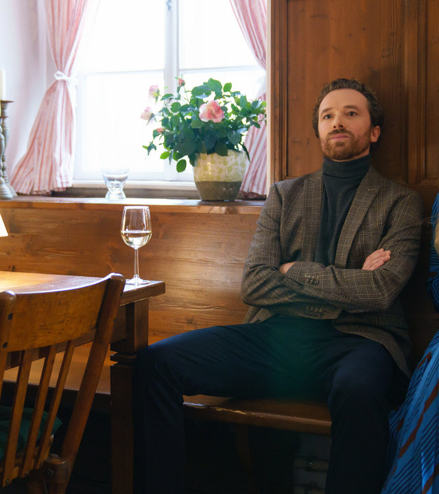 "SOKO Wien -  Mein Essen mit Erik": Auf einer Holzbank in einem Restaurant sitzen Penny Lanz (Lilian Klebow) und Erik Maurer (Philip Kelz) nebeneinander. Vor Ihnen auf dem Tisch steht ein Weinglas. Penny Lanz hält ihres in der rechten Hand.