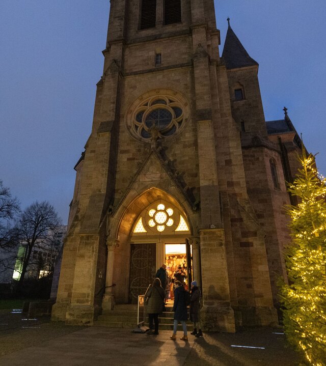 "Evangelische Christvesper - Wonach suchst Du?": Bei der Christuskirche in Fulda ist am Heiligabend die Tür geöffnet und lädt zum Eintreten ein. Zwei beleuchtete und festlich geschmückte Weihnachtsbäume stehen links und rechts vor der Kirche.
