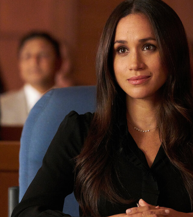 "Suits - Schande": Rachel (Meghan Markle) sitzt schwarz gekleidet mit gefalteten Händen und selbstsicherem Blick an einem Tisch.  Im Hintergrund sind in Sitzbänken Zuschauer zu erkennen.