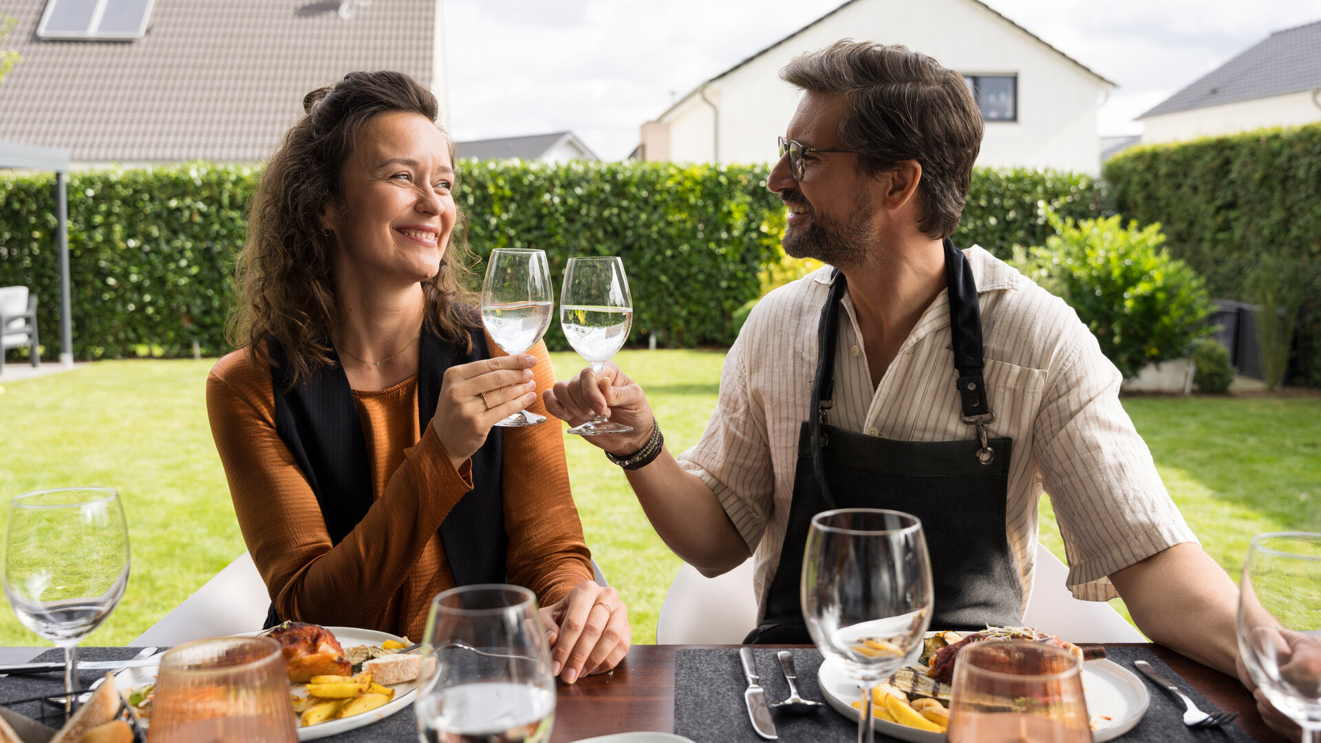 "Like a Loser: Ernst macht auf ernst": Marie (Tinka Fürst) und Guideon (Tom Beck) sitzen gemeinsam an einem gedeckten Tisch im Garten. Beide heben lächelnd ihre Weingläser zum Anstoßen und schauen sich dabei strahlend an. Vor ihnen stehen Teller mit gegrilltem Essen, Wein- und Wassergläser.