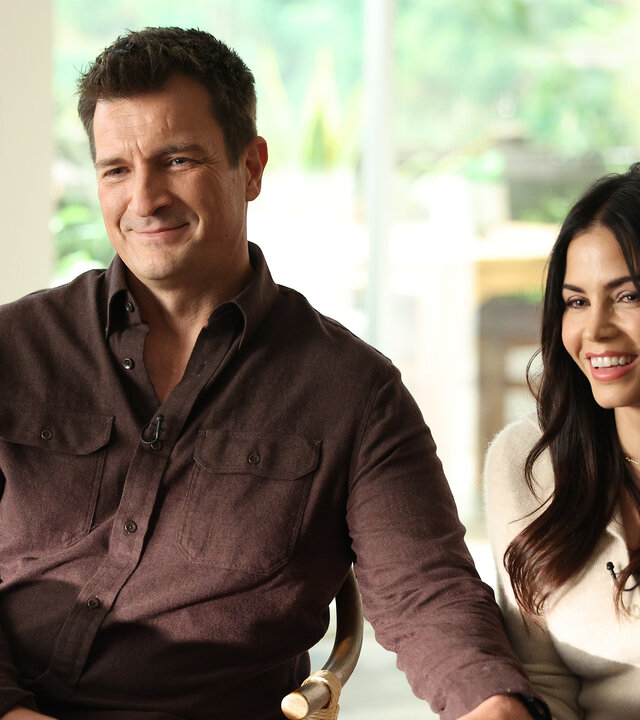 "The Rookie - Double Trouble": John Nolan (Nathan Fillion) und Bailey (Jenna Dewan) sitzen im Wohnzimmer Seite an Seite und lächeln den Interviewer an, während ihre Hände liebevoll ineinander verschränkt sind.