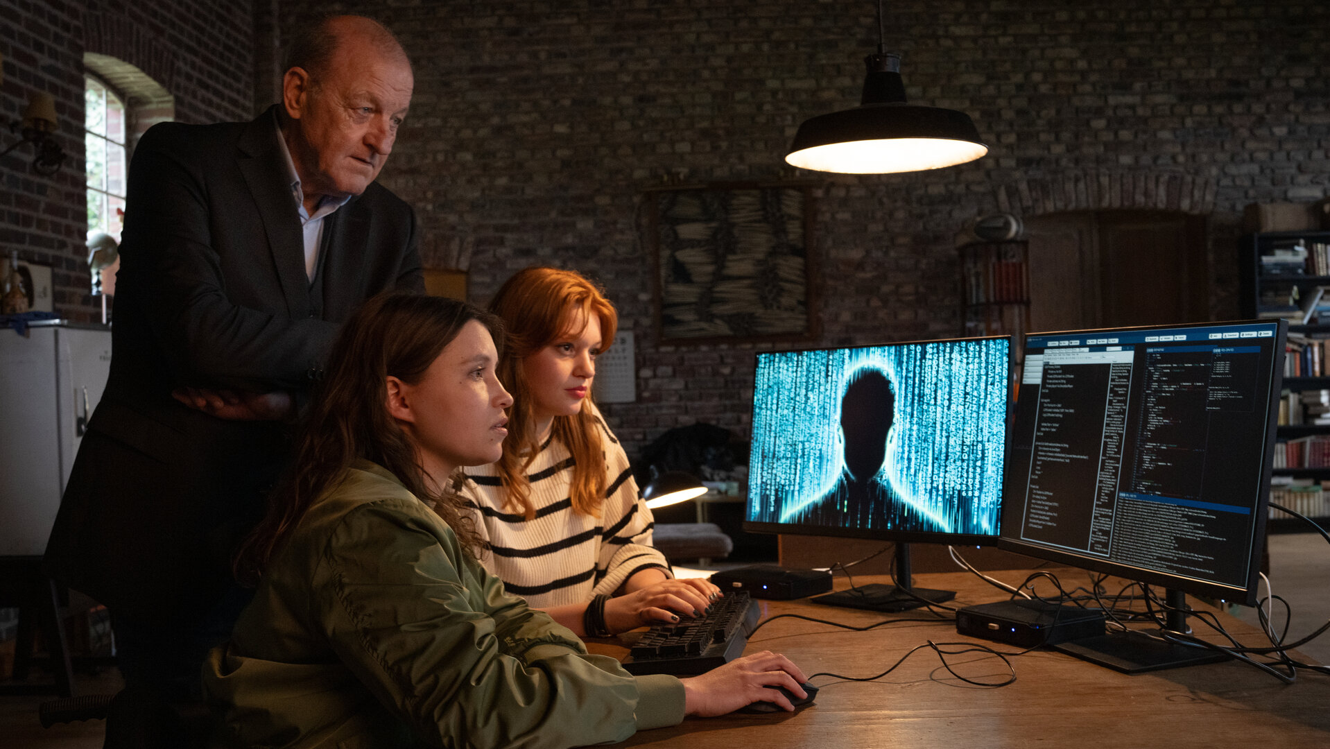 "Wilsberg - Phantomtod": Jana Haumann (Lilly Charlotte Dreesen) und Merle (Janina Fautz) sitzen an einem Tisch und bedienen eine Computer-Maus und eine Tastatur, während sie gebannt auf die beiden Bildschirme vor sich starren. Wilsberg (Leonard Lansink) steht hinter ihnen und blickt ebenfalls auf die Monitore, auf denen unter anderem eine dunkle Silhouette einer Person zu sehen ist.