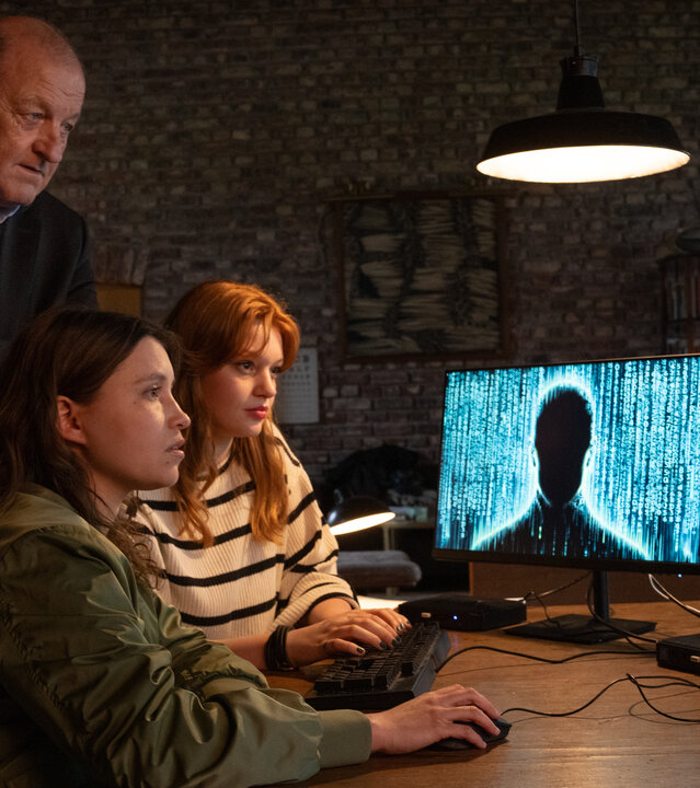 "Wilsberg - Phantomtod": Jana Haumann (Lilly Charlotte Dreesen) und Merle (Janina Fautz) sitzen an einem Tisch und bedienen eine Computer-Maus und eine Tastatur, während sie gebannt auf die beiden Bildschirme vor sich starren. Wilsberg (Leonard Lansink) steht hinter ihnen und blickt ebenfalls auf die Monitore, auf denen unter anderem eine dunkle Silhouette einer Person zu sehen ist.